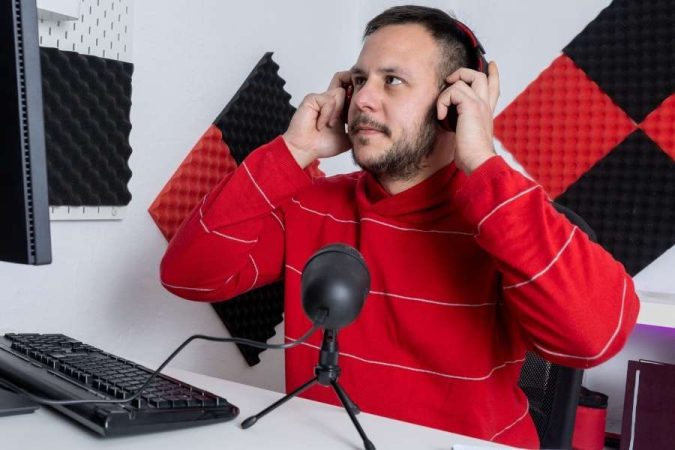 Un homme dans un studio d'enregistrement portant des écouteurs et regardant un moniteur, avec un microphone et une insonorisation murale.