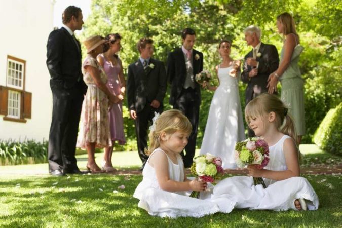 Deux adorables petites filles d'honneur en robe blanche, assises sur une pelouse verte, jouent avec des bouquets de fleurs lors d'une cérémonie de mariage en plein air.